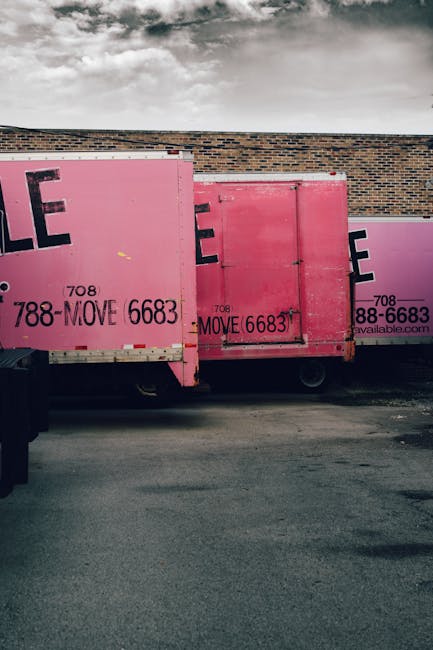 An image showing three large pink moving containers positioned side by side along a brick wall exterior. The containers are made of metal with printed black text and contact details, and each features a compartment with a door for storing packed belongings. The containers are placed on a paved surface, and part of a black vehicle is visible at the left edge. The site appears to be prepared for a home relocation or furniture transport process managed by Man with Van Belmont. The containers are outdoors under a cloudy sky, ready for loading or unloading, which aligns with typical moving and packing activities involved in professional removals services.