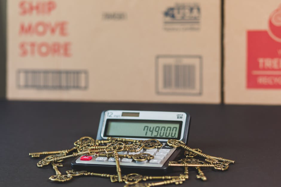 Close-up view of a small calculator displaying the number 749800, surrounded by scattered antique brass keys, some attached to small chains, positioned on a dark surface. In the background, there are large cardboard boxes with printed labels indicating shipping and recycling symbols, placed in an indoor environment that suggests a packing or moving process. The scene captures elements involved in home relocation, highlighting the preparatory packaging phase and the calculation of moving costs, which relates to professional removals services offered by Man with Van Belmont. The lighting is even, emphasizing the details of the keys and the calculator, with the boxes slightly out of focus to maintain focus on the objects in the foreground.