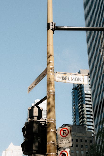 A street scene showing a tall beige utility pole with multiple metal bands, situated at an urban intersection under a clear blue sky. Attached to the pole are various street signs, including a white sign displaying 'RUE BELMONT' and another indicating 'Robert F. Bourdais'. Partially visible traffic lights are mounted on an arm extending from the pole, and additional signage below indicates parking restrictions, such as no parking during certain hours. Surrounding the pole are modern high-rise buildings with glass facades, and the street environment suggests a busy city area. This setting, associated with Belmont, relates to house removals and moving logistics, as part of a typical relocation or furniture transport process involving urban streets and city infrastructure, which Man with Van Belmont may assist with during home relocation services.