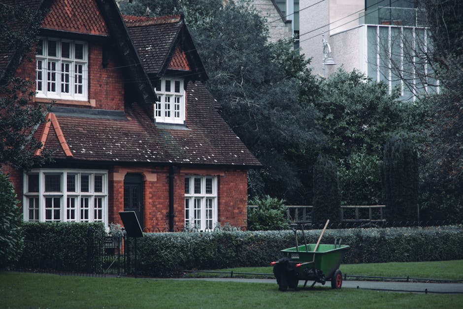 Outside a traditional red-brick house with white-framed sash windows and a dark tiled pitched roof, a green wheelbarrow with black handles and red wheels is positioned on the paved driveway in front of a neatly trimmed hedge. Inside the wheelbarrow, there are two wooden sticks, one topped with a small plastic item, and the other appears to have a metal hook. Nearby, a black toolbox and a small black bag rest on the pavement, suggesting a packing or loading activity associated with home relocations. The setting is during daylight hours with natural lighting, and nearby trees and shrubbery create a leafy environment. The background includes a modern multi-storey building, indicating the house is likely situated in an urban or suburban area. This scene depicts the process of packing and moving small garden items as part of a house removal service, with equipment ready for transporting household or garden belongings, aligning with the contents discussed on the Belmont Park to new home -- small-garden items removal guide page by Man with Van Belmont.