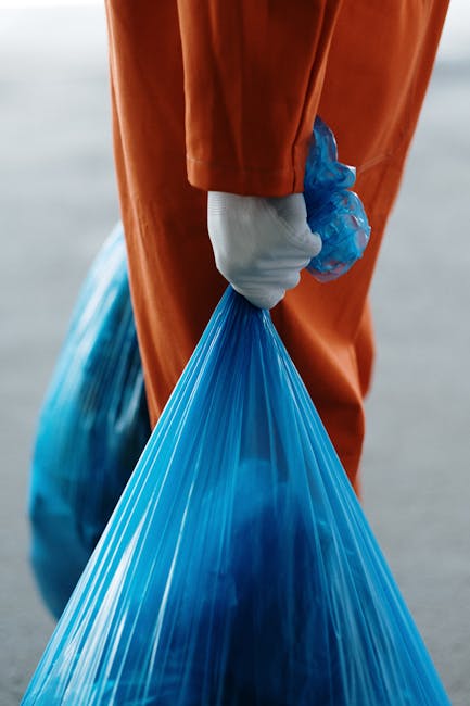 A close-up of a person wearing orange protective overalls and white gloves, holding a large blue plastic waste bag filled with rubbish. The individual is standing outdoors on a pavement or driveway, with some blurred background elements. The focus is on the action of waste disposal, which may relate to household or commercial waste management during a home relocation or moving process. The image highlights the packing and decluttering aspect of house removals, with the bag visibly filled with waste materials, supporting themes of waste disposal and local council regulations as discussed in Belmont removals' recent guidelines. Man with Van Belmont offers services that include managing waste disposal as part of their comprehensive moving and furniture transport solutions, ensuring compliance with local regulations. The scene emphasizes safe handling of waste and organized packing during moving logistics, with the practical action of waste collection conveying efficiency and care in relocation tasks.