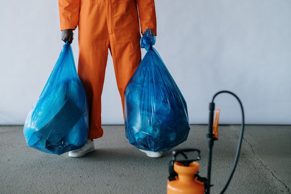 A person dressed in orange work overalls, with white shoes, is holding two large blue plastic rubbish bags filled with waste, standing on a gray floor inside a neutral-colored room. The person's face and upper body are not visible, only their lower torso and legs. In the foreground, there is an orange completely visible vacuum cleaner or carpet extractor with a black hose attachment, situated on the floor. This scene depicts the process of waste disposal as part of house removal or relocation activities, which could include packing or clearing during a home move. The background features a plain light-colored wall, emphasizing the indoor environment typical of a house or storage facility, and the lighting is neutral. The image relates naturally to moving logistics, packing, and waste disposal procedures often necessary during a relocation or professional removals service by companies like Man with Van Belmont.
