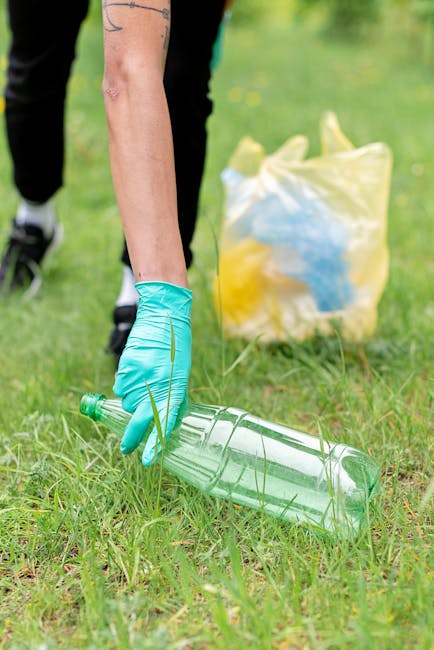 A close-up of a person wearing orange protective overalls and white gloves, holding a large blue plastic waste bag filled with rubbish. The individual is standing outdoors on a pavement or driveway, with some blurred background elements. The focus is on the action of waste disposal, which may relate to household or commercial waste management during a home relocation or moving process. The image highlights the packing and decluttering aspect of house removals, with the bag visibly filled with waste materials, supporting themes of waste disposal and local council regulations as discussed in Belmont removals' recent guidelines. Man with Van Belmont offers services that include managing waste disposal as part of their comprehensive moving and furniture transport solutions, ensuring compliance with local regulations. The scene emphasizes safe handling of waste and organized packing during moving logistics, with the practical action of waste collection conveying efficiency and care in relocation tasks.