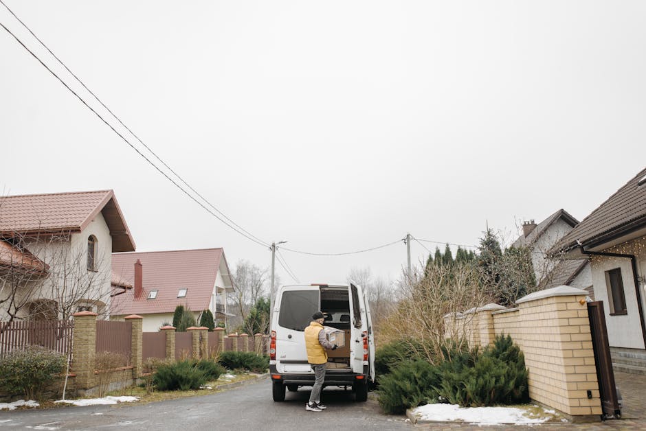 A man dressed in a red and black uniform stands at the rear of a white van, loading or unloading items during a home relocation. The van is parked on a paved driveway outside a modern two-story house 