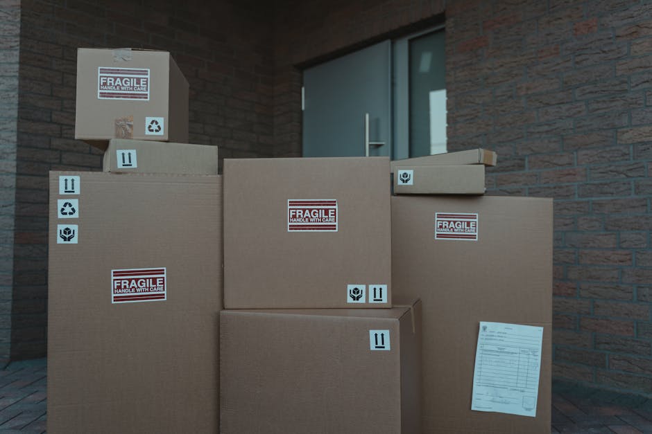 A collection of cardboard boxes of various sizes stacked outside a residential property, with some boxes marked with 'fragile handle with care' labels and others with symbols indicating careful handling and orientation instructions. The boxes are positioned on a paved surface near a brick wall and a modern door with a glass panel, suggesting they are in the process of being moved or prepared for home relocation. The scene appears to depict furniture transport or packing materials used during a house removal, with a focus on the organized loading process managed by Man with Van Belmont, a professional removals service specializing in household moves and logistics.