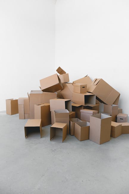 A large pile of various-sized cardboard boxes, some flattened and others still assembled, situated in a corner of a white room with a plain white wall and grey floor. The boxes are made of brown cardboard, with some open and others closed, and a few show visible tape or packing labels. The boxes appear to be part of a home relocation process, possibly unpacked or awaiting packing as part of furniture transport and removals. No furniture or additional packaging materials are visible, and the lighting is even, highlighting the cluttered arrangement typical of unpacking or packing during a house move. This scene reflects the logistical aspects of home removals managed by services such as Man with Van Belmont, featuring typical packing materials used in moving and transport processes.