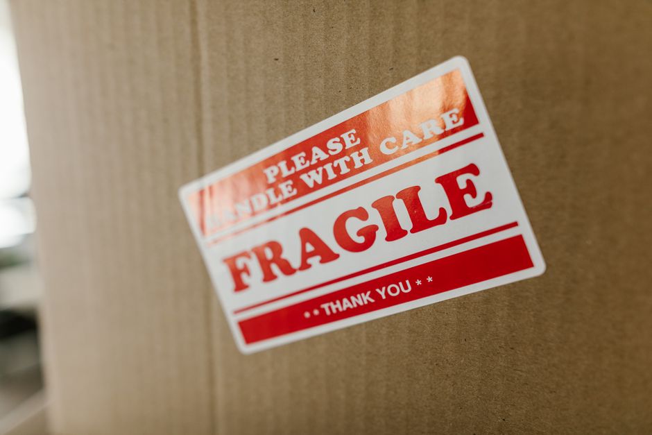 A collection of cardboard boxes of various sizes stacked outside a residential property, with some boxes marked with 'fragile handle with care' labels and others with symbols indicating careful handling and orientation instructions. The boxes are positioned on a paved surface near a brick wall and a modern door with a glass panel, suggesting they are in the process of being moved or prepared for home relocation. The scene appears to depict furniture transport or packing materials used during a house removal, with a focus on the organized loading process managed by Man with Van Belmont, a professional removals service specializing in household moves and logistics.