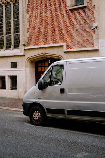 A grey commercial van is parked on the street in front of a historic red-brick building with large stone window surrounds and small windows near the ground. The van is positioned close to a doorway with an arched stone lintel leading into the building. The side door of the van is visible, and the vehicle is situated on a paved road with no other cars in sight. This setting illustrates typical home relocation or furniture transport logistics, which may involve parking permits for removal vans. The environment is well-lit with natural light, and the scene represents a clean, urban residential area where professional house removals by companies like Man with Van Belmont typically operate during moving and packing processes.