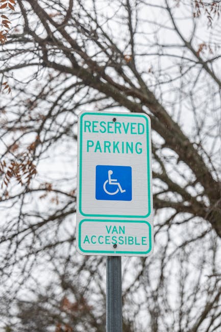 A vertical rectangular signpost mounted on a metal pole, displaying a white background with green and blue text and symbols. The top section reads 'RESERVED PARKING' in green uppercase letters, accompanied by a blue wheelchair symbol indicating disabled parking. Below, an additional section states 'VAN ACCESSIBLE' in green letters, confirming suitable parking for removals vans. The sign is situated outdoors against a backdrop of leafless tree branches and a cloudy sky, suggesting an urban or suburban street environment. This signage relates to parking regulations often encountered during home relocation and furniture transport services, as provided by companies like Man with Van Belmont, who facilitate efficient packing and loading processes within permit-controlled areas.