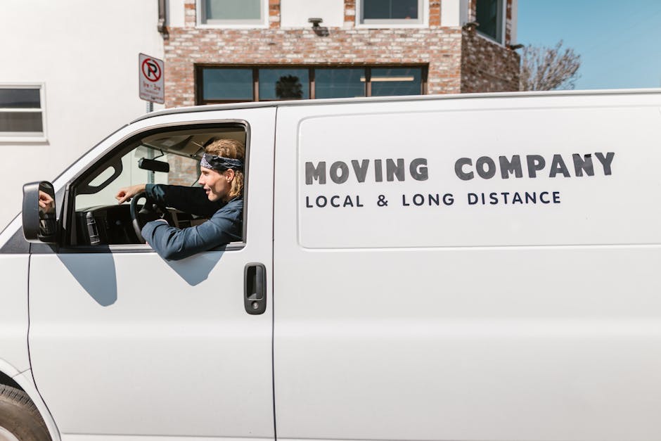 A white moving van parked on a street near a brick residential building with large windows, with the company's logo 'MOVING COMPANY, LOCAL & LONG DISTANCE' printed on its side. Inside the open driver's door, a male mover with shoulder-length blond hair, wearing a black headband, a dark blue shirt, and black gloves, is shown loading the vehicle. He is holding a cardboard box wrapped in protective plastic, preparing to lift it onto the van. The scene includes a paved sidewalk, a pavement parking restriction sign, and clear daylight, illustrating a home relocation or furniture transport scene connected to professional removals services. The image reflects the logistical process of packing and loading for a house move near Belmont Station, with Man with Van Belmont specializing in house removals and moving coordination.