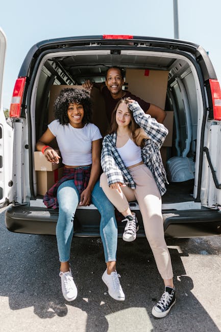Three individuals, two young women and one man, are seated inside the open rear of a black moving van, preparing for a home relocation. The woman on the left has curly black hair with a light dusting of snow-like material, wearing a white t-shirt and light blue jeans, with her right arm resting on the van's threshold. The woman on the right has long straight hair and is dressed in a white top and beige trousers, with a checkered shirt tied around her waist, and is adjusting her hair. The man standing behind them, with short dark hair and a broad smile, wears a dark t-shirt and jeans, and appears to be helping with the loading process. The interior of the van shows large boxes made of cardboard and wrapped with plastic or fabric for protection, some stacked behind the seated women. The environment is outdoors on a paved surface with bright, natural lighting suggesting daytime, and the focus is on the furniture and packing materials ready for transport, supporting professional removals and furniture transport services by Man with Van Belmont.
