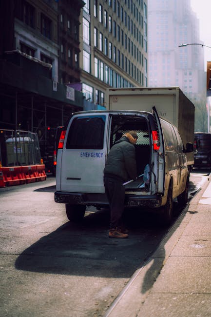 A person wearing a dark jacket and beige hat loading or unloading items from the back of a white emergency van parked on a city street. The van's rear doors are open, revealing the cargo space with boxes and packing materials, possibly plastic wrap or fabric, inside. The van is positioned next to a curb, with orange and black safety barriers on the sidewalk nearby. Surrounding buildings are tall and modern, with reflective glass windows, and other vehicles are parked along the street. The scene is lit by natural daylight, indicating daytime, and the environment suggests a busy urban area suitable for residential or commercial removals and home relocation processes, with Man with Van Belmont providing furniture transport and packing services.