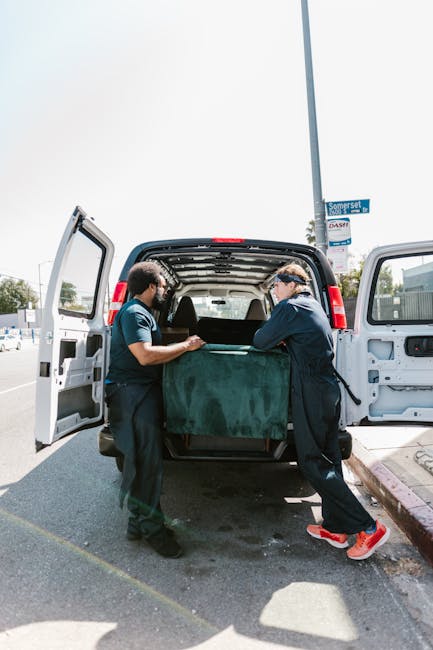 Two movers from Man with Van Belmont are loading a large piece of furniture, covered with a green moving blanket, into the rear cargo space of a black van parked on the street in Belmont. The interior of the van is visible with its back doors open, revealing the spacious loading area. One mover, dressed in dark clothing and wearing safety glasses, supports the bottom of the furniture while the other, also dressed in dark clothing with red and grey sneakers, helps lift the item. The scene takes place outdoors on a clear day, with a streetlamp and some trees visible in the background, alongside a street sign that reads 'Somerset.' The moving process appears carefully managed, reflecting home relocation activities and furniture transport involved in a house removal or packing and moving operation, exemplifying the sort of service provided by Man with Van Belmont for urgent or same-day moves.