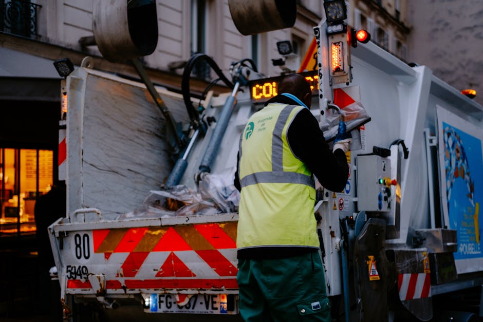 A worker wearing a neon yellow high-visibility vest and dark clothing is operating a waste collection vehicle in an urban setting during dusk or evening. The vehicle has a large open rear compartment filled with discarded bulky waste such as cardboard boxes, plastic-wrapped items, and wooden furniture. The worker is seen manipulating controls on the side of the vehicle, possibly preparing to unload or collect waste, with their back to the camera. The collection vehicle is marked with red and white reflective strips and features an electronic display showing 'CO1'. The scene is illuminated by ambient urban lighting and the vehicle's internal lights, emphasizing the process of home clearance or removal of bulky waste items, aligned with the services provided by Man with Van Belmont. The background includes a building with windows emitting warm light, indicating an evening schedule for waste collection in connection with house removals or clutter clearance services.