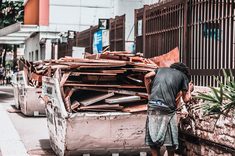A man with curly hair, dressed in a dark T-shirt and checkered shorts, stands on a sidewalk next to a large open skip filled with various building waste materials such as wooden planks, cardboard, and debris, which are stacked inside the container. The skip is positioned outdoors along a street, with a metal fence and some greenery visible on the right side of the image. In the background, there are other skips and a few pedestrians, along with a building façade featuring window signs and a partially visible canopy. The scene reflects a waste clearance or house removal process, where materials are being prepared for disposal or recycling, consistent with services offered by Man with Van Belmont for bulky waste removal and home relocation assistance.